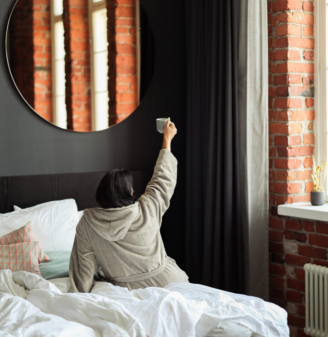 Syksyn kampanja hotellihuoneista – Folks Hotels Person in a gray robe sits on a bed, raising a cup toward a round mirror on a dark wall with brick walls and curtains in view behind.