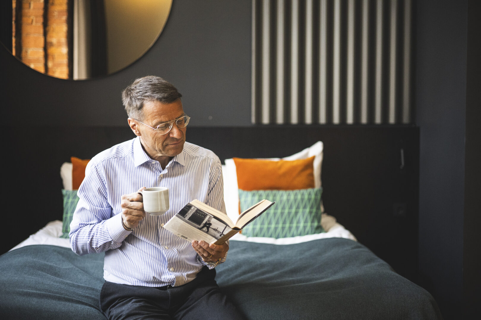 Older man with glasses sitting on a bed, reading a newspaper and holding a cup in a cozy room with orange and green cushions behind him
