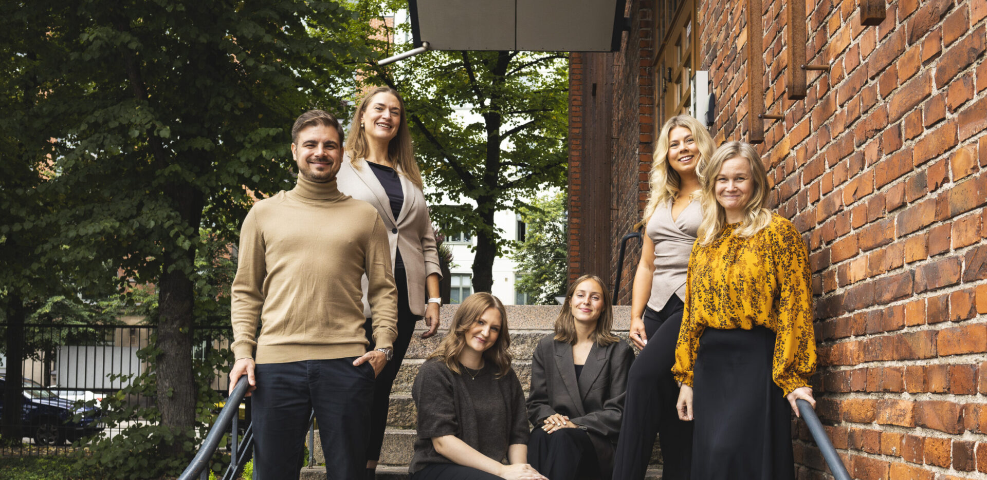 Folks_henksu_2025 – Folks Hotels Six adults posing on outdoor stone steps beside a brick wall, with trees in the background, smiling at the camera.